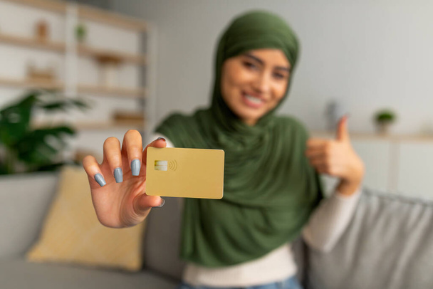 Feliz joven árabe sosteniendo tarjeta de crédito y mostrando gesto de pulgar hacia arriba en casa, enfoque selectivo - Foto, imagen