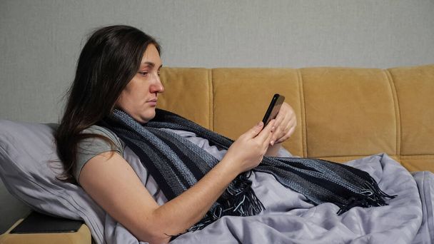 Mujer enferma se sienta en el sofá y ordena medicamentos a través de un teléfono inteligente - Foto, Imagen