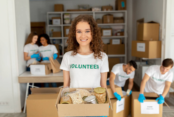 Positivo Millennial Lady volontario in possesso di scatola di donazione di cibo e sorridente alla macchina fotografica, lavorando presso l'organizzazione di beneficenza - Foto, immagini