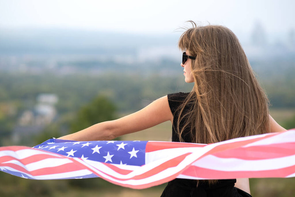 Young pretty american woman with long hair holding waving on wind USA flag on her sholders resting outdoors enjoying warm summer day. - Photo, Image