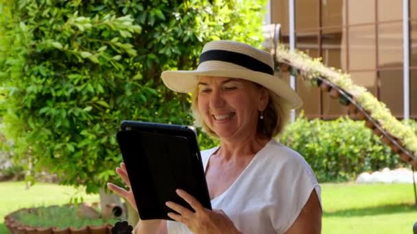 gerencia Senior mujer rubia empresaria en sombrero utiliza tableta PC, tableta digital para el trabajo de negocios o estudio en su propio jardín verde. mujer de 50 a 55 años. El concepto de su propia empresa y formación - Metraje, vídeo