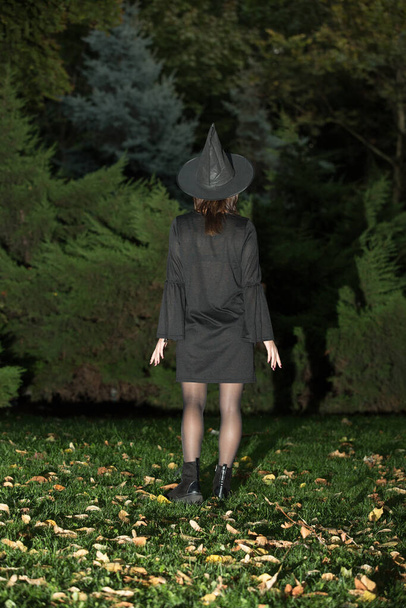 A woman in a black hat and black dress in full growth with her back to the camera. Fashionable girl in retro style. Black hat with large brim. Halloween witch. - Foto, imagen