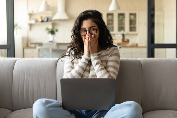 Junge kaukasische Frau schaut auf Laptop-Bildschirm, lacht, bedeckt ihren Mund, schaut lustige Filme am PC zu Hause - Foto, Bild
