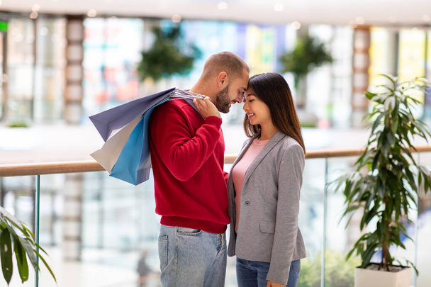 Στοργικό νεαρό ζευγάρι με φωτεινές τσάντες δώρων στέκεται, αγγίζοντας τα μέτωπα, εκφράζοντας την αγάπη στο εμπορικό κέντρο της πόλης - Φωτογραφία, εικόνα
