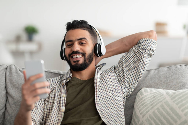 Пора расслабиться. Заручний молодий арабський чоловік у бездротових навушниках слухає музику в Інтернеті на смартфоні вдома. - Фото, зображення