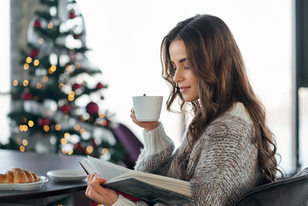 Young woman reading book near Christmas tree at home. - Foto, Imagen