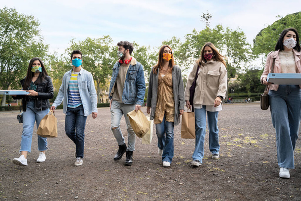 Un grupo de jóvenes caminan por el parque y usan máscaras protectoras, llevándose paquetes de comida para llevar con ellos para pasar un domingo juntos - Foto, Imagen