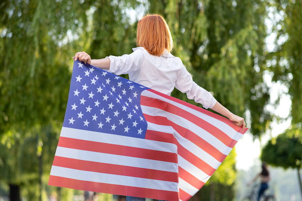 Οπίσθια όψη χαρούμενης νεαρής κοκκινομάλλας γυναίκας που ποζάρει με την εθνική σημαία των ΗΠΑ να στέκεται σε εξωτερικούς χώρους στο πάρκο. Θετικό κορίτσι γιορτάζει την ημέρα ανεξαρτησίας των Ηνωμένων Πολιτειών. - Φωτογραφία, εικόνα