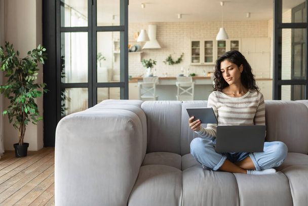 Молода кавказька пані, користуючись ноутбуком і цифровим планшетом, вивчаючи або працюючи в Інтернеті, сидячи на дивані вдома, копіюйте простір. - Фото, зображення