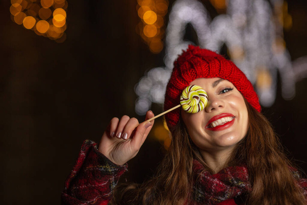 Όμορφη νεαρή γυναίκα με κόκκινο καπέλο που κρατάει γλυκά γλυκά σε χριστουγεννιάτικο πανηγύρι. Χώρος για κείμενο - Φωτογραφία, εικόνα