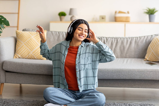 Positive young Arab woman sitting on floor with headphones, listening to music or audio book at home - 写真・画像