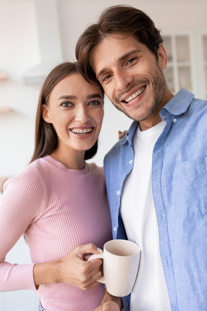 Portret van vrolijke glimlachende jonge vrouw en echtgenoot met kopje warme drank fotograferen op de keuken - Foto, afbeelding