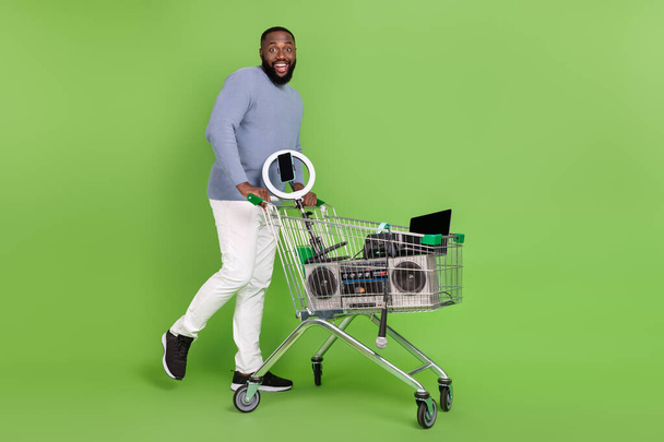 Photo of shocked afro guy wear pullover walking shopping tray modern devices isolated green color background - Zdjęcie, obraz