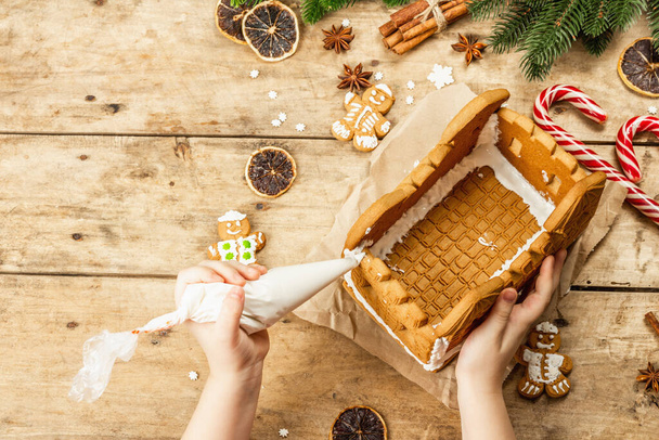 Το παιδικό χέρι διακοσμεί το Gingerbread House με γλυκό κερασάκι. Χειροποίητη διαδικασία, εορταστική διακόσμηση Πρωτοχρονιάς. Παραδοσιακά μπαχαρικά, ξύλινο φόντο, top view - Φωτογραφία, εικόνα