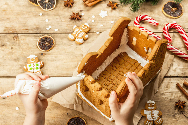 La main des enfants décorer Gingerbread House par glaçage doux. Processus fait à la main, décor festif du Nouvel An. Épices traditionnelles, fond bois, gros plan - Photo, image