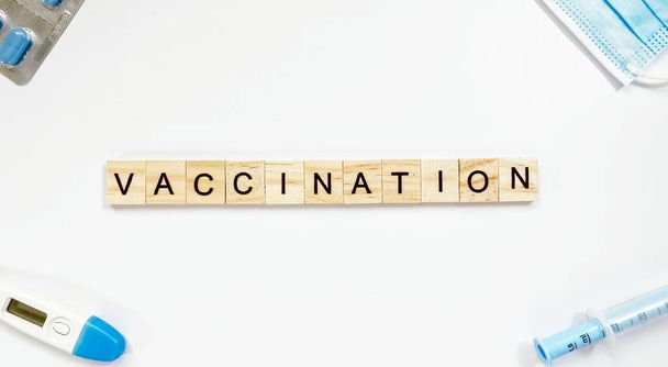 Макет со словом вакцинация из деревянных букв и рядом со шприцем, термометром, лекарственной маской и таблетками - Фото, изображение