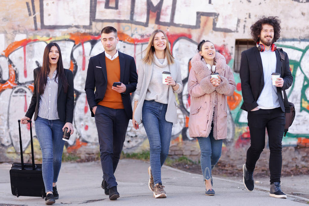 Group of people walking on a street with confidence. Businessmen and businesswomen traveling together. Old wall with abstract unrecognizable graffiti in the background. - Фото, изображение