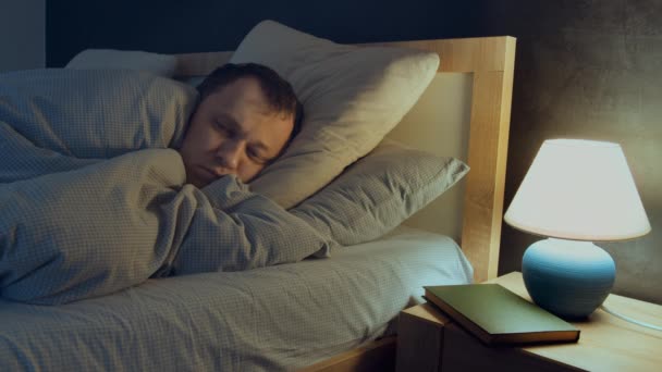 Człowiek ma bezsenność, nie może zasnąć leżąc w nocy w łóżku, lampa jest na stoliku nocnym - Materiał filmowy, wideo
