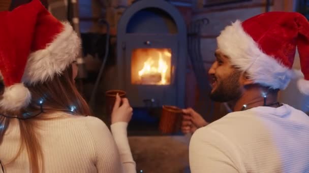 Pareja cariñosa con sombreros de santa bebiendo bebida caliente cerca de la chimenea en la víspera de Navidad - Metraje, vídeo