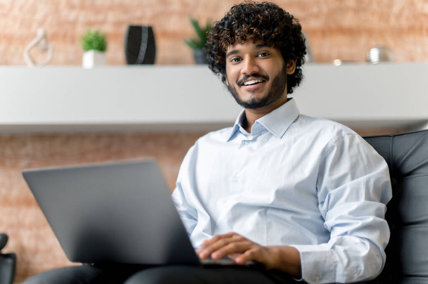 Indian positive confident curly-haired young adult guy, freelancer or manager in shirt, sitting on armchair with laptop looking at the camera and smiling friendly - Fotografie, Obrázek
