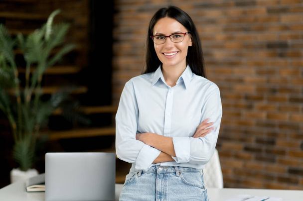 Portrait of successful confident pretty young adult caucasian businesswoman or broker, stand near work desk in office, wearing formal stylish clothes, arms crossed, looking at camera, smiles friendly - Zdjęcie, obraz