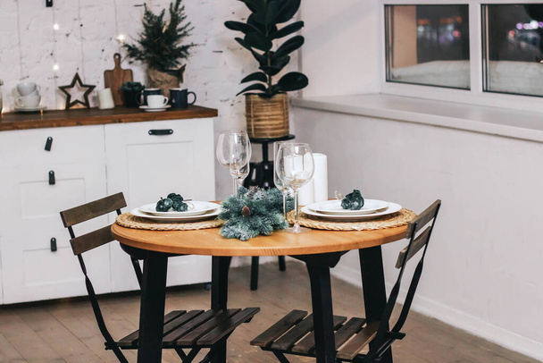 Round table decorated for Christmas and New Year. Christmas tree twigs on the table. Plates and wine glasses on the table. Kitchen set and flowers on the background. Christmas content. - Foto, Imagem