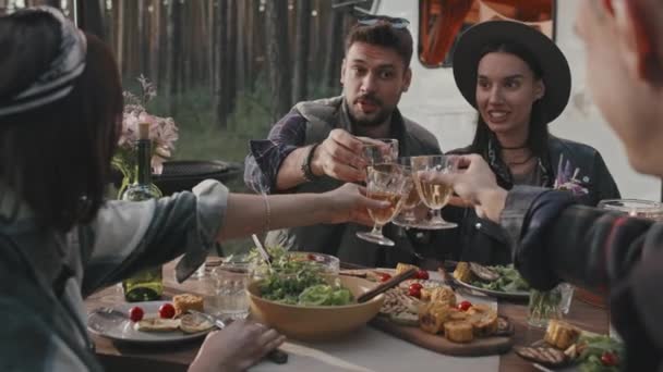 Handheld slow mo van gelukkige jonge mannen en vrouwen klinkende hun glazen tijdens het diner op de camping op de zomeravond. Witte camper geparkeerd op de achtergrond - Video
