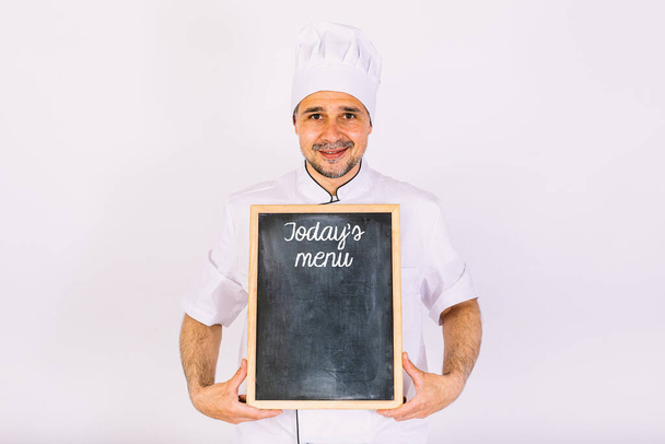 Chef kok in jas en keukenhoed glimlachend, met een bord met het menu, op witte achtergrond - Foto, afbeelding