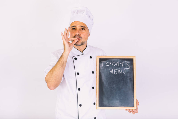 Chef cocinero en chaqueta y sombrero de cocina sonriendo, sosteniendo una tabla con el menú, sobre fondo blanco - Foto, imagen