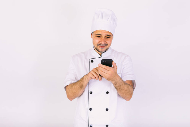 Cook vêtu d'un chapeau blanc et d'une veste regardant son téléphone portable sur fond blanc. Concept restaurant, cuisine et plats à emporter. - Photo, image