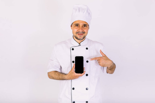 Fehér kalapba és kabátba öltözött séf, fehér alapon mutatja a mobiltelefonját. Étterem, étel és elvitel koncepciója. - Fotó, kép