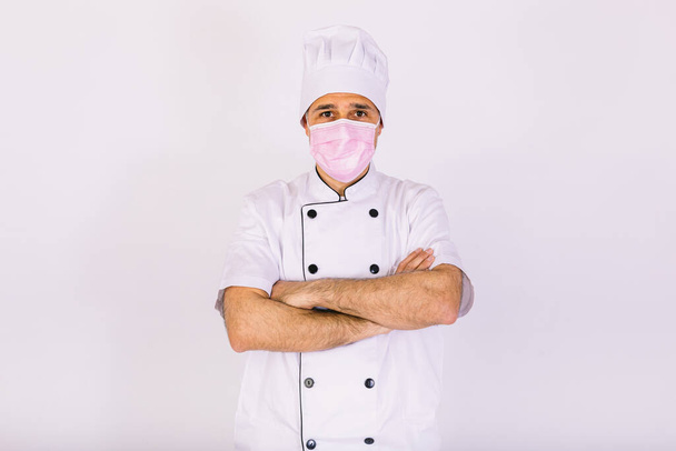 Chef kok draagt keukenjas en hoed, met roze masker, kruisende armen, op witte achtergrond - Foto, afbeelding