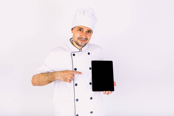 Chef cuisinier vêtu d'une veste et d'un chapeau de cuisine, tenant une tablette, montrant son écran, sur fond blanc - Photo, image