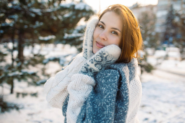Портрет рыжеволосой девушки в варежках на фоне зимнего парка. Красивая женщина наслаждается природой в снежный день - Фото, изображение