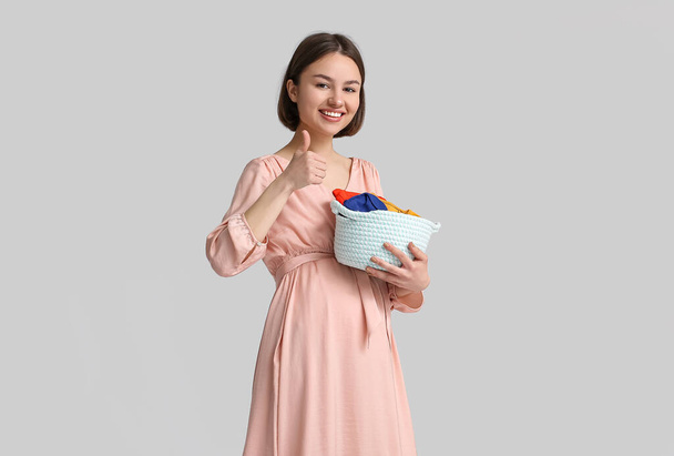 Young pregnant woman holding basket with baby clothes on light background - Foto, afbeelding