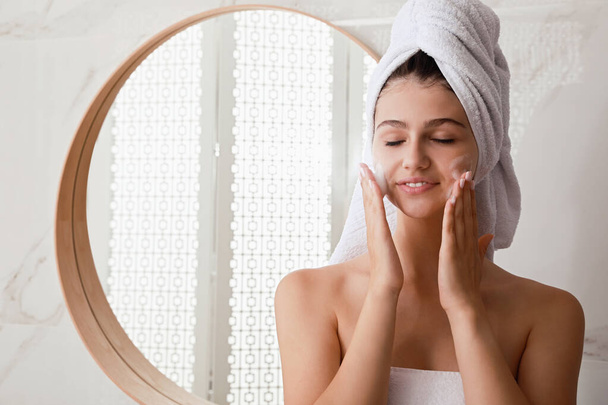 Beautiful teenage girl applying cleansing foam onto face in bathroom, space for text. Skin care cosmetic - Фото, зображення