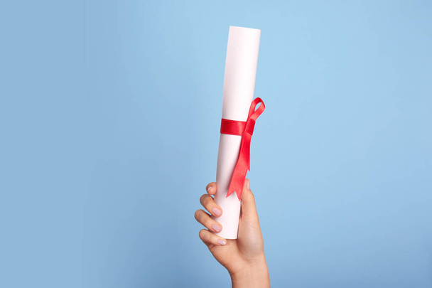 Student holding rolled diploma with red ribbon on light blue background, closeup - Photo, image