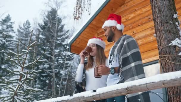 Noel sabahı. Dağdaki ahşap evin önünde karla kaplı sıcak bir içecek içen mutlu genç çift. - Video, Çekim