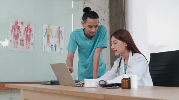 Egészségügyi csapattársak vagyunk. Két egyenruhás, fiatal ázsiai etnikai orvos kollégák, akik gyógyszerekről beszélgetnek a kórház rendelőjében. A szakemberek szakértők és szakemberek.. - Felvétel, videó