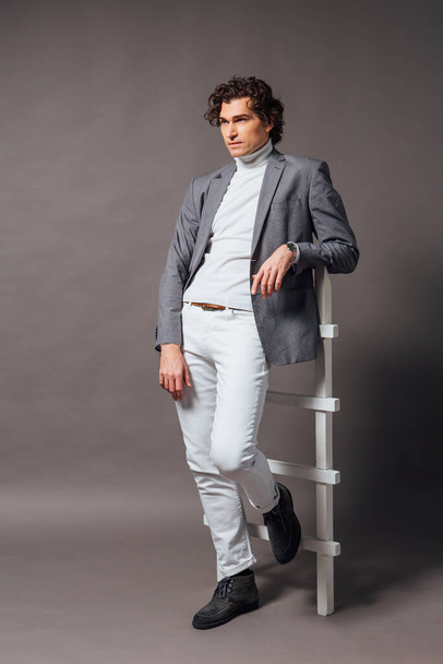 Fashion portrait of a tall handsome man dressed in white turtleneck, white jeans and grey jacket posing on the grey background with white ladder - Fotoğraf, Görsel