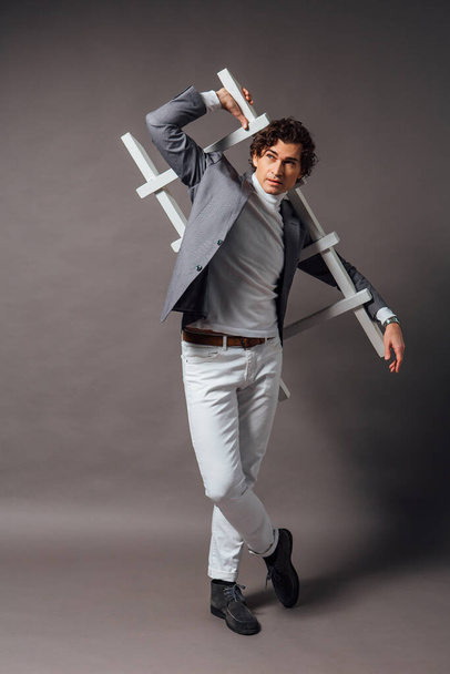 Fashion portrait of a tall handsome man dressed in white turtleneck, white jeans and grey jacket posing on the grey background with white ladder - Foto, afbeelding