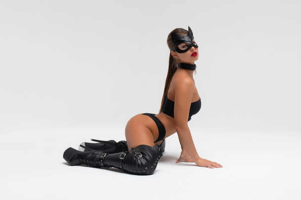 Full body side view of alluring slim tanned brunette in black mask and underwear with high heeled leather boots in white studio - Фото, зображення