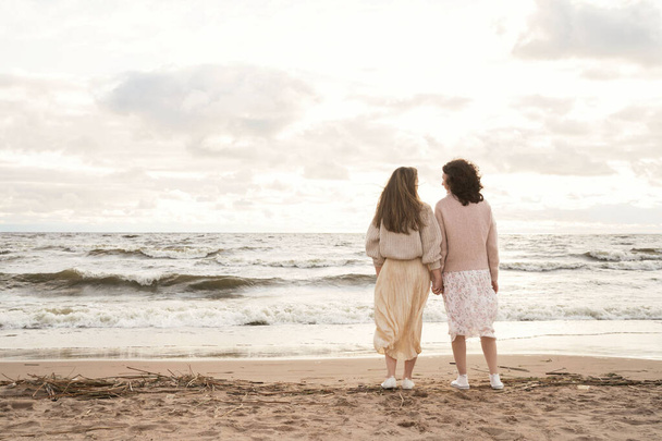 Δύο κορίτσια πάνε από το χέρι στη θάλασσα. Περπατήστε δίπλα στον ωκεανό το φθινόπωρο με ζεστά ρούχα. Σχέσεις μεταξύ γυναικών. - Φωτογραφία, εικόνα