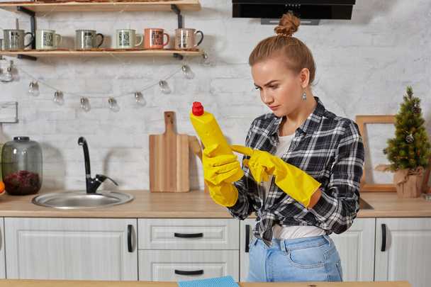 Professional young woman wearing rubber protective yellow gloves holding bottle cleaners in the kitchen. - 写真・画像