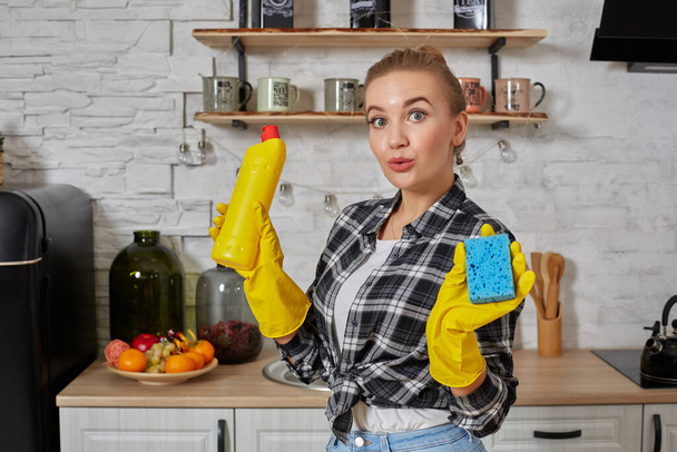 Profesjonalna młoda kobieta nosi gumowe rękawice ochronne żółte trzymając butelki czyszczące w kuchni. - Zdjęcie, obraz