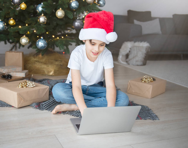 A boy sits near a Christmas tree with a laptop. Christmas tree in a large living room. - Photo, Image