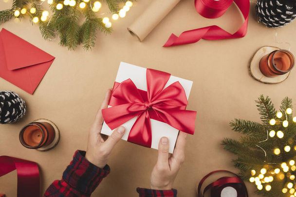 Kırmızı kurdeleli beyaz hediye kutusu Kraft arka planında el ele tutuşuyor. Kutlama için hediye paketleme ve hazırlama. Yılbaşı arkaplanı ağaç süsü ve ışıklarıyla - Fotoğraf, Görsel
