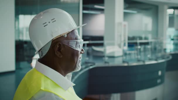 Ingénieur masculin noir en vêtements de protection debout à l'intérieur au centre d'affaires moderne - Séquence, vidéo
