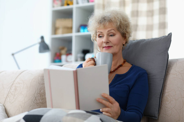 Az idős hölgy könyvet olvas és kávét iszik, hogy otthon kényelmesen legyen a kanapén. - Fotó, kép
