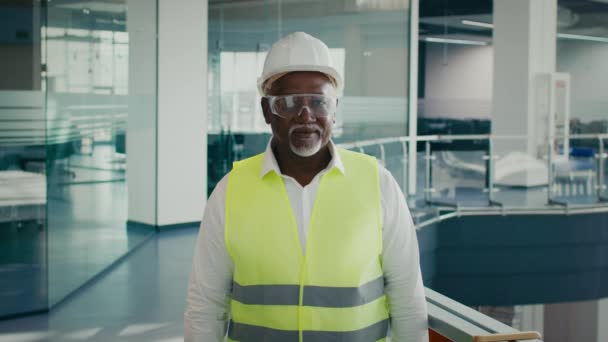 Kypsä musta rakennusinsinööri suojavaatetuksessa poseeraa tyhjässä liikekeskuksessa - Materiaali, video
